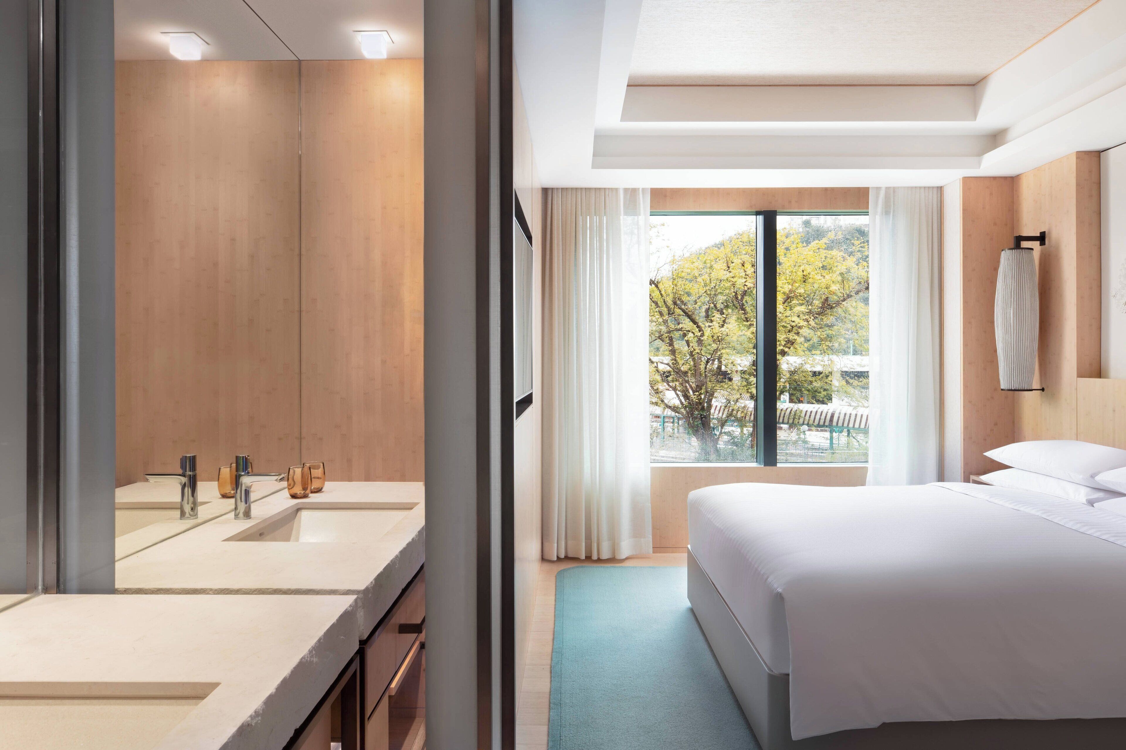 Ein Hotelzimmer mit großem Spiegel, einem Bett, einem Fenster mit Vorhängen und Blick auf Bäume.