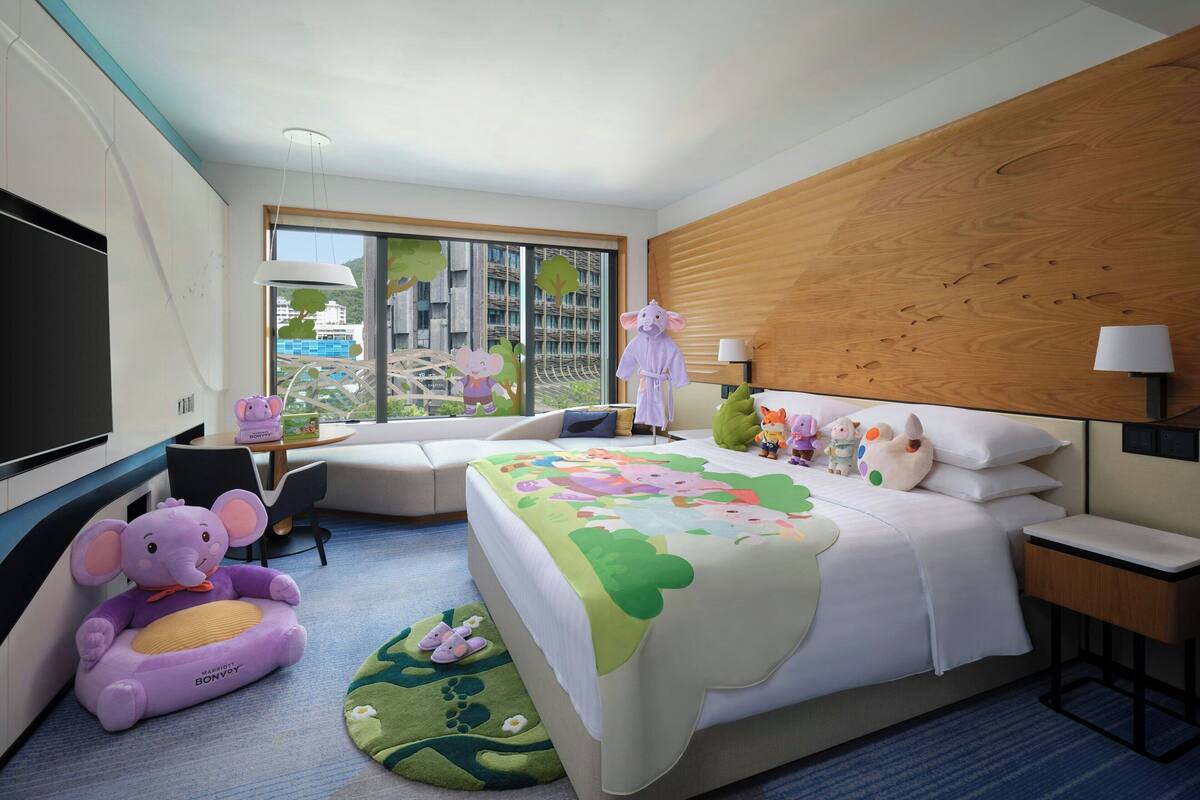 A hotel room with a large bed, a desk, a chair, a TV, and a window with a view of buildings.