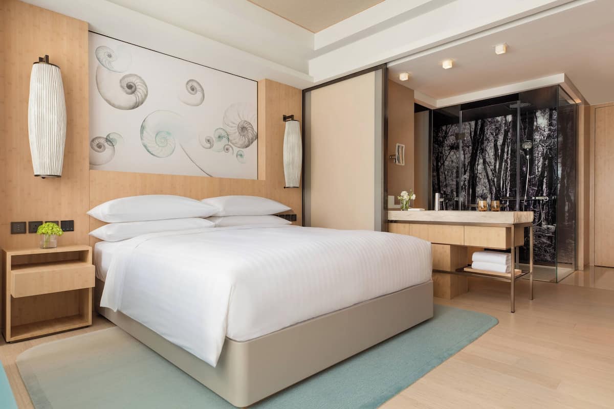 A modern bedroom with a large bed, a bedside table with a vase, a wall-mounted artwork, and a bathroom with a large mirror and a sink.