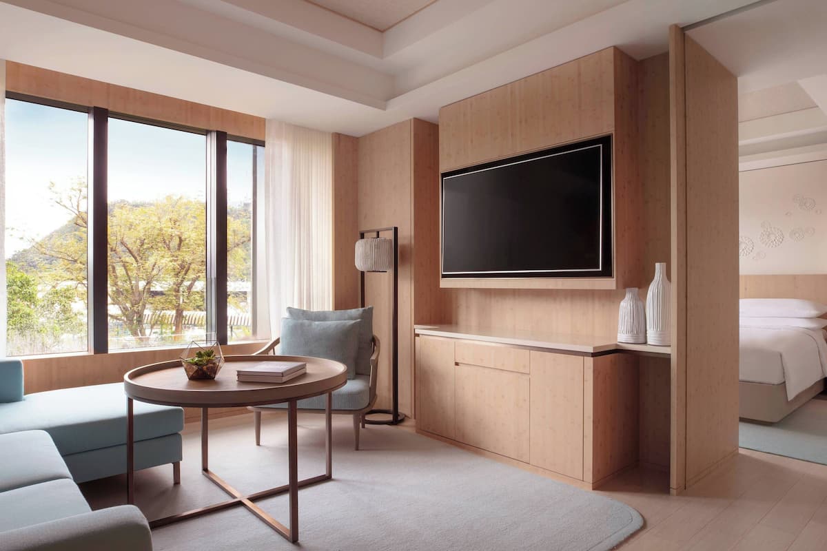 A modern hotel room with a large window, a flat-screen TV, a sofa, a round table, and a bed.