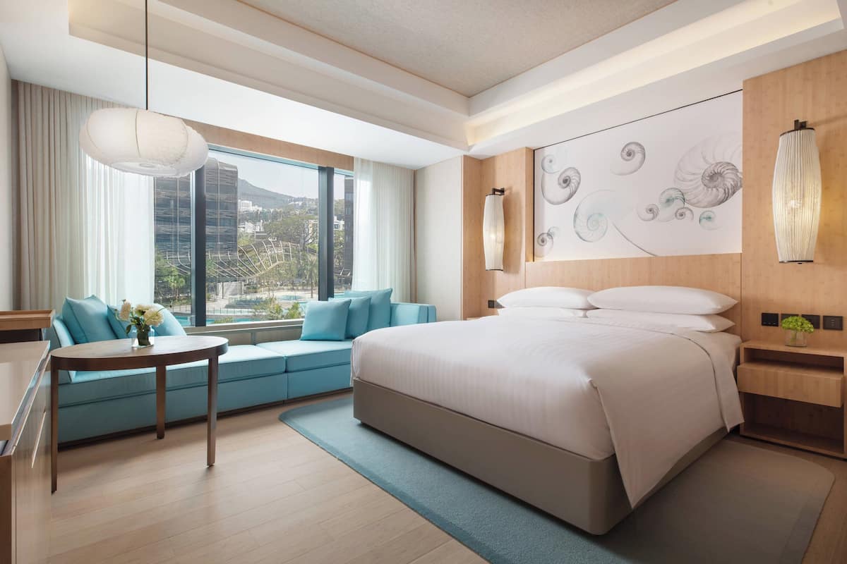 A modern hotel room with a large bed, a sofa, a dining table, and a view of the cityscape through large windows.