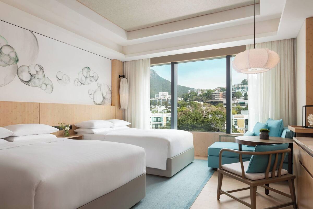 A hotel room with two beds, a small table with a chair, a large window with a view of buildings and greenery, and a ceiling with a light fixture.
