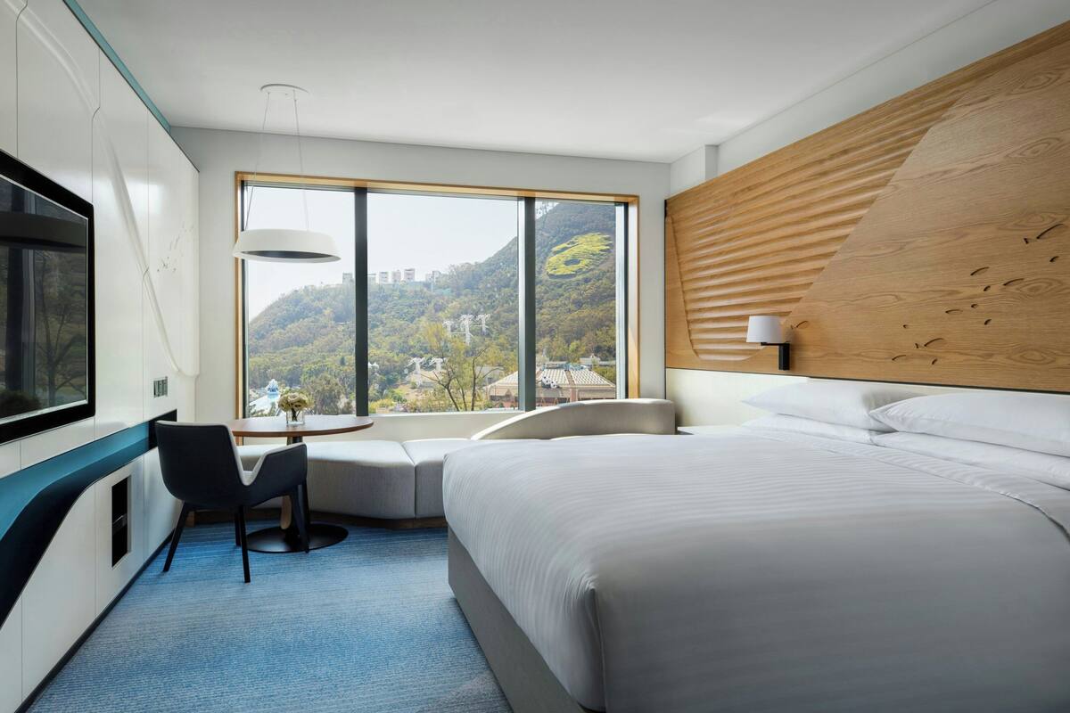 A modern hotel room with a large bed, a seating area with a table, a TV, and a view of a mountainous landscape.
