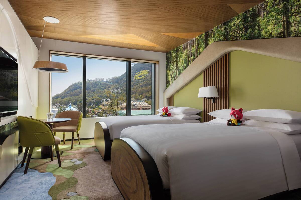A hotel room with two beds, a dining area with chairs, a TV, and a large window offering a view of a mountainous landscape.