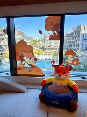 Un peluche sobre un cojín, una ventana con vistas a edificios y una piscina, y un panel decorativo con un personaje de caricatura.