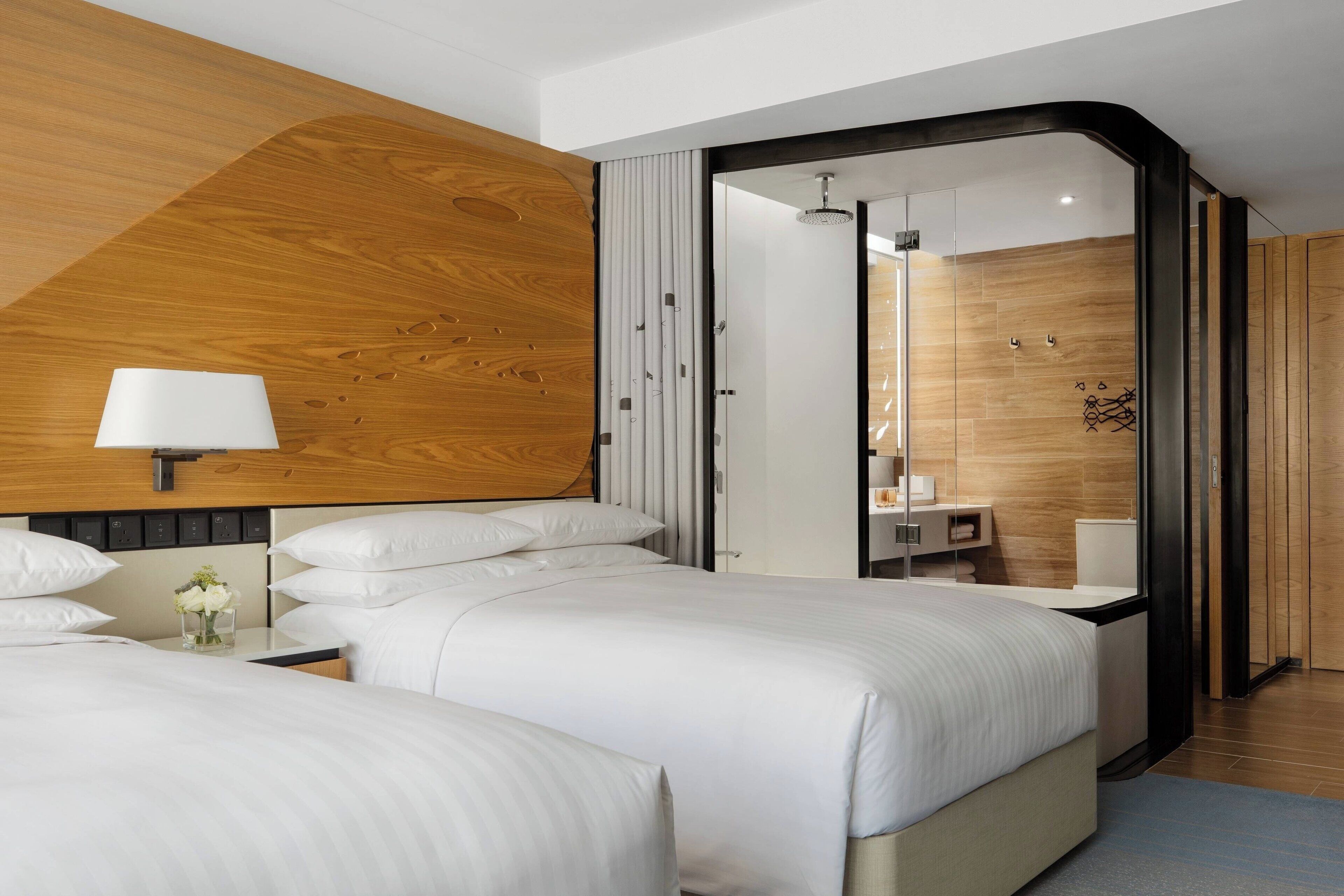 Habitación de hotel moderna con dos camas, un panel de madera decorativo, cabina de ducha de vidrio y una mesita de noche con lámpara.