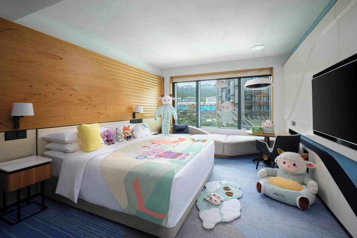 A hotel room with a large bed, a TV, a desk, and a window with a view of buildings.