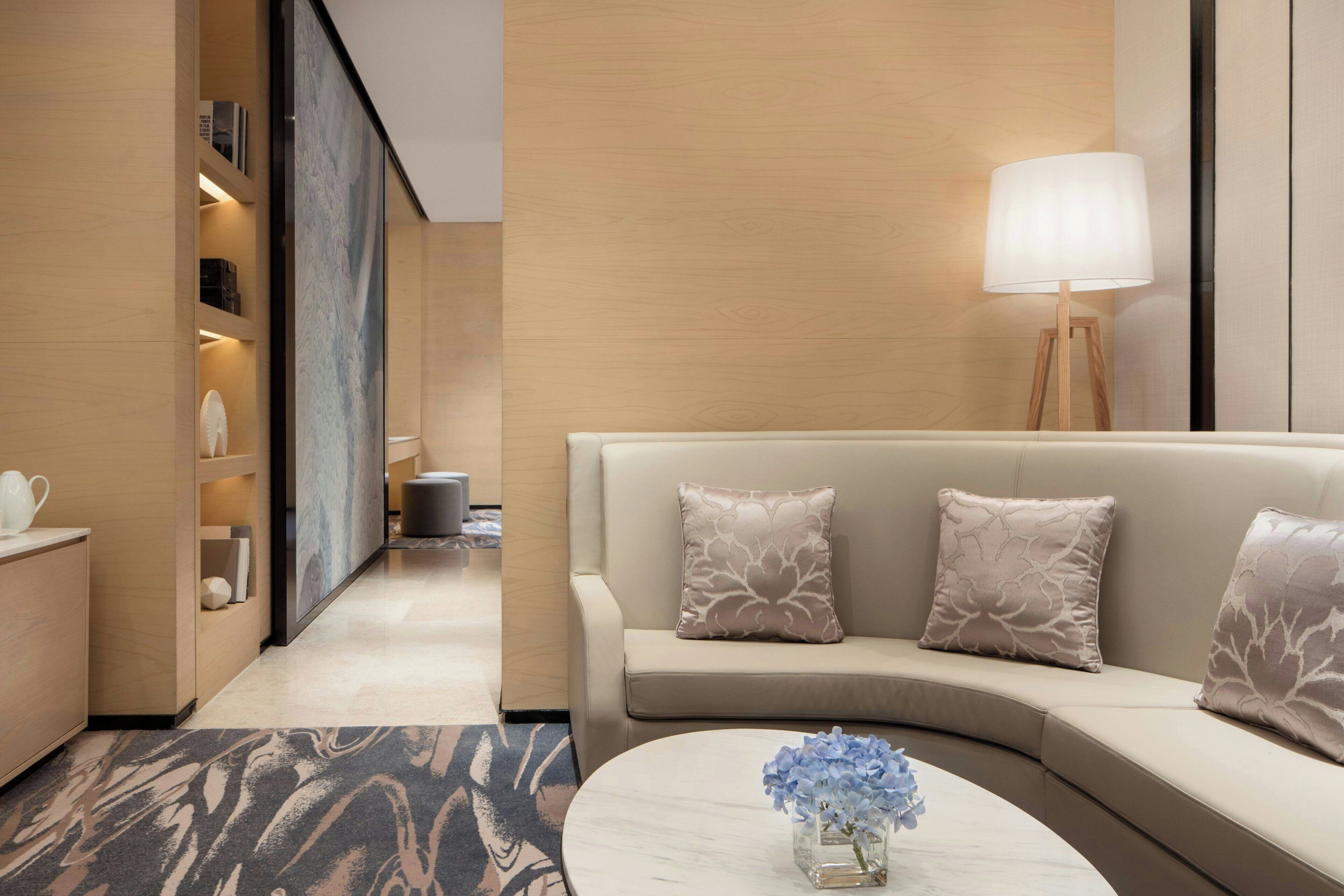 Et moderne hotelværelse med en buet sofa, et lille sofabord med en vase blomster, en lampe og udsigt over bybilledet gennem et stort vindue.