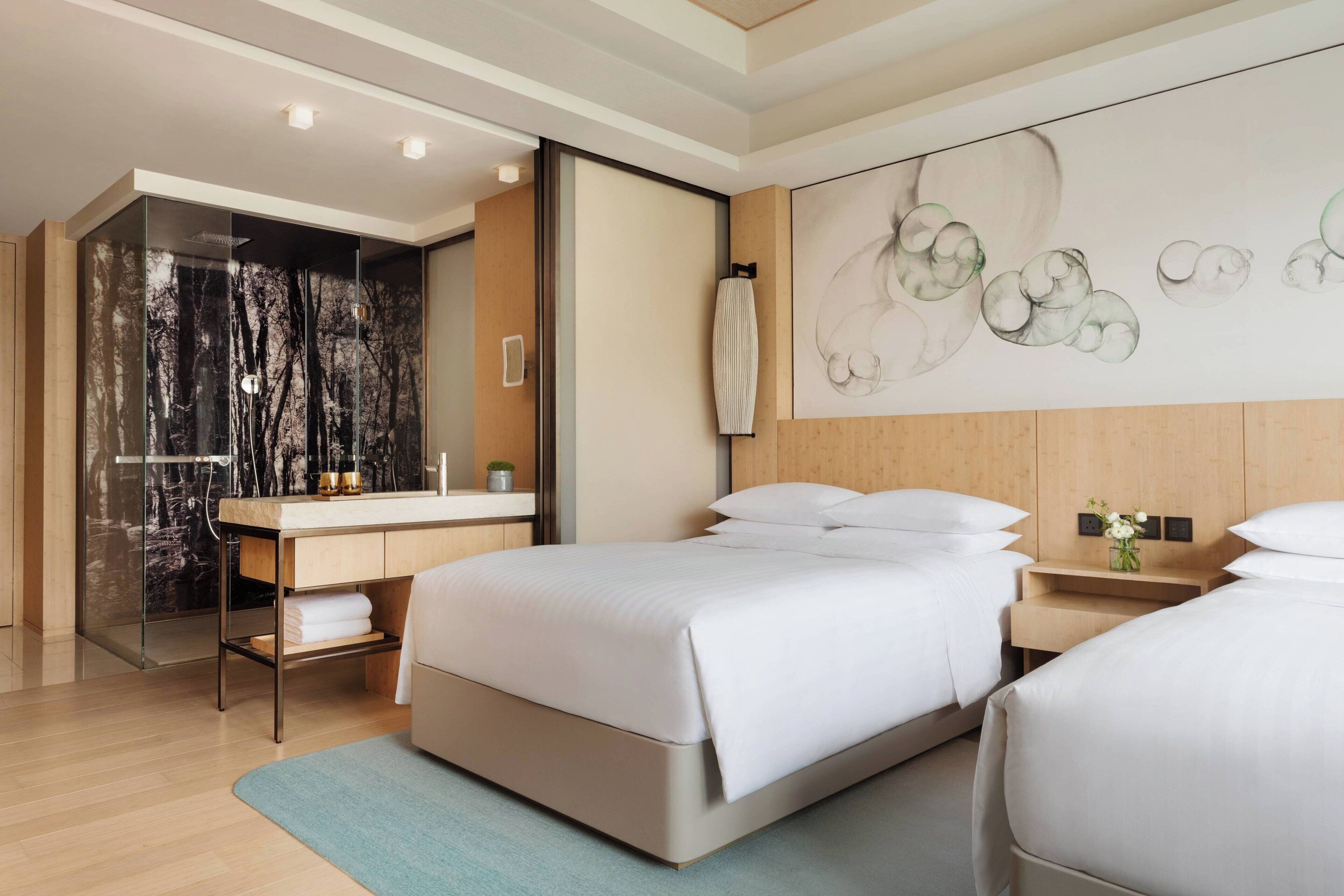 A modern hotel room with a large bed, a separate bathroom with a glass shower, a wooden headboard, and a wall art piece.