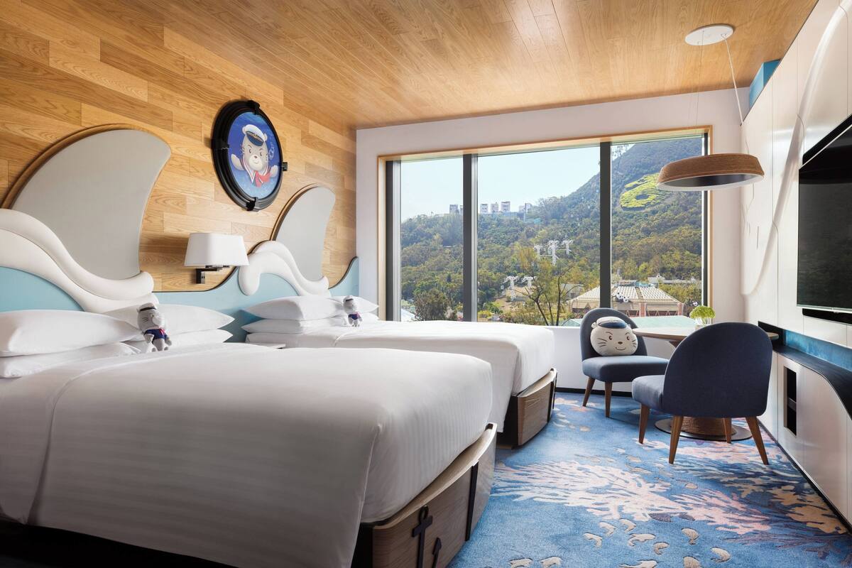A hotel room with two beds, a large window with a mountain view, a desk with a chair, and a wall-mounted TV.