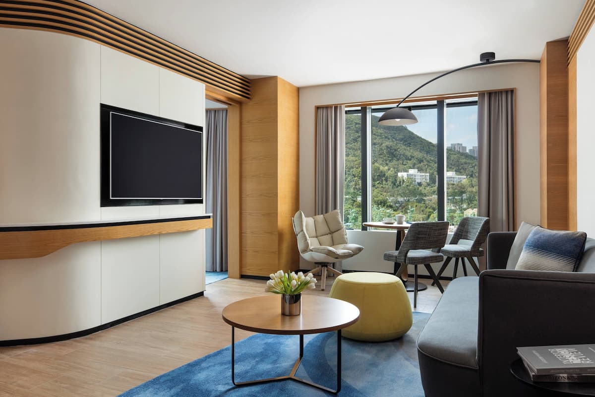 A modern hotel room with a flat-screen TV, comfortable seating, a large window with a mountain view, and a blue carpet.