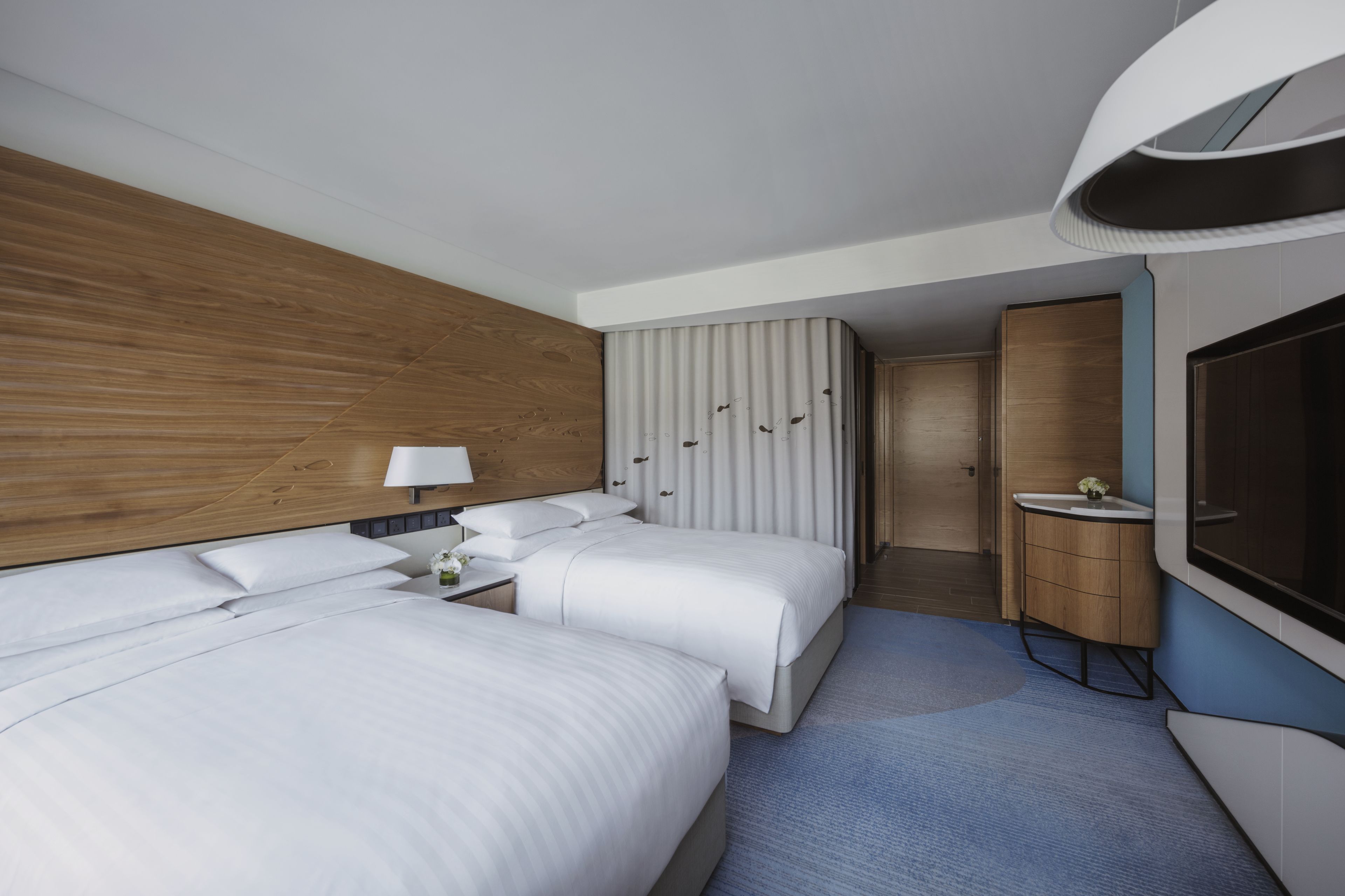 Ein modernes Hotelzimmer mit zwei Betten, einem Holz-Kopfteil, einem großen Spiegel, einem Fernseher und einer kleinen Badeinrichtung.
