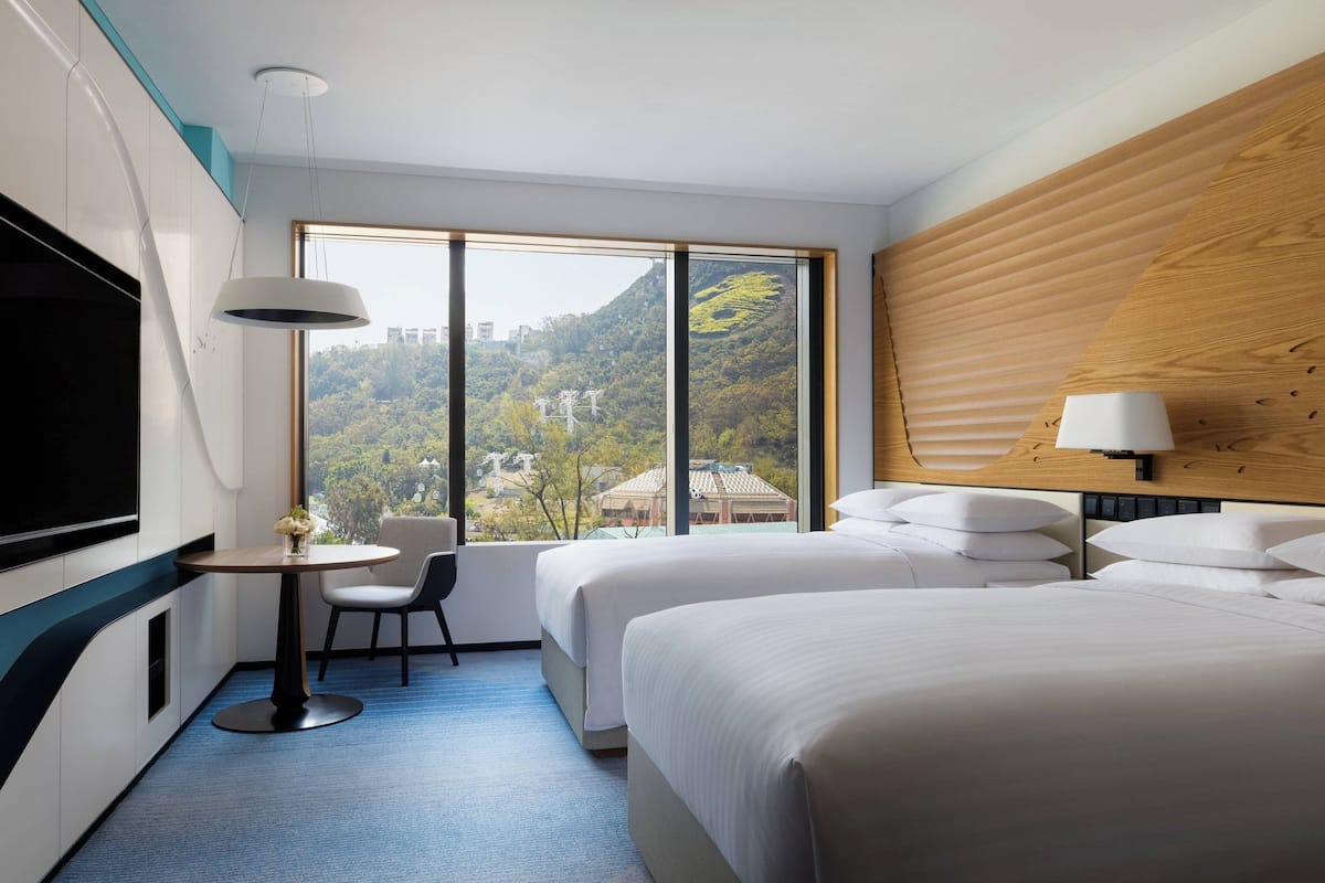 A hotel room with two beds, a TV, a small table with a chair, and a large window offering a view of a mountainous landscape.