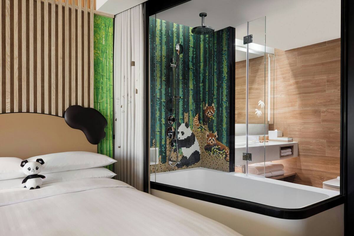 A modern bedroom with a bed, a plush panda toy, a sliding glass door leading to a bathroom with a mosaic wall, a shower, and a toilet.