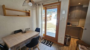 A dining area with a table and chairs, a hanging light fixture, a view through a door, and a small trash can. - Valmorel - Cozy apartment for 4, direct access to slopes (Valmorel)