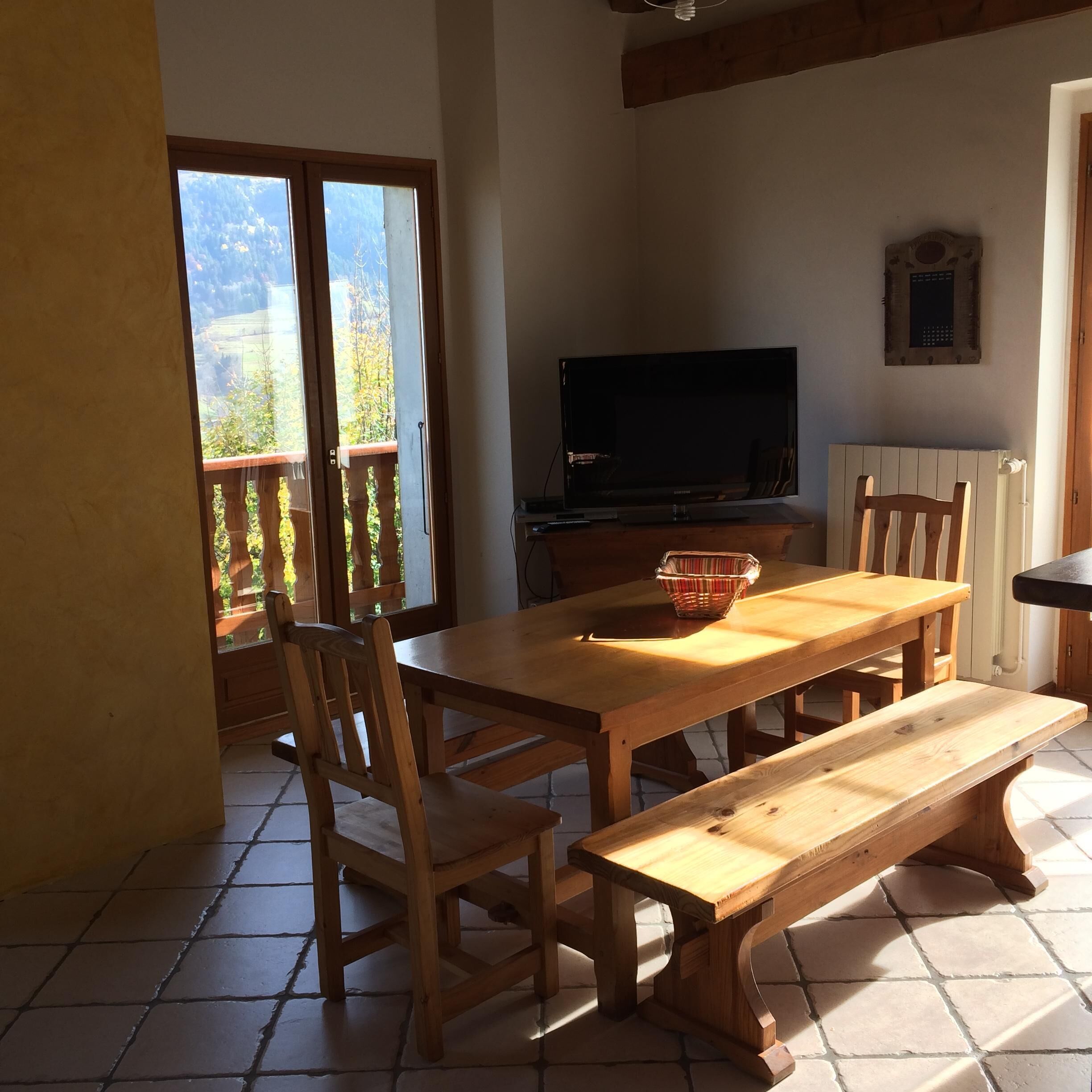 Zona pranzo con tavolo e panche in legno, televisore su un mobile e vista all'esterno tramite una porta scorrevole.