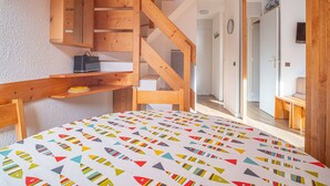 Ein Schlafzimmer mit einer Holztreppe, einem Bett mit farbenfroher geometrischer Bettdecke und einem kleinen Holzregal mit einer Tasche und einem Buch.