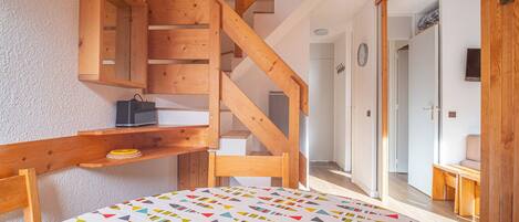 A bedroom with a wooden staircase, a bed with a colorful geometric-patterned bedspread, and a small wooden shelf with a bag and a book.