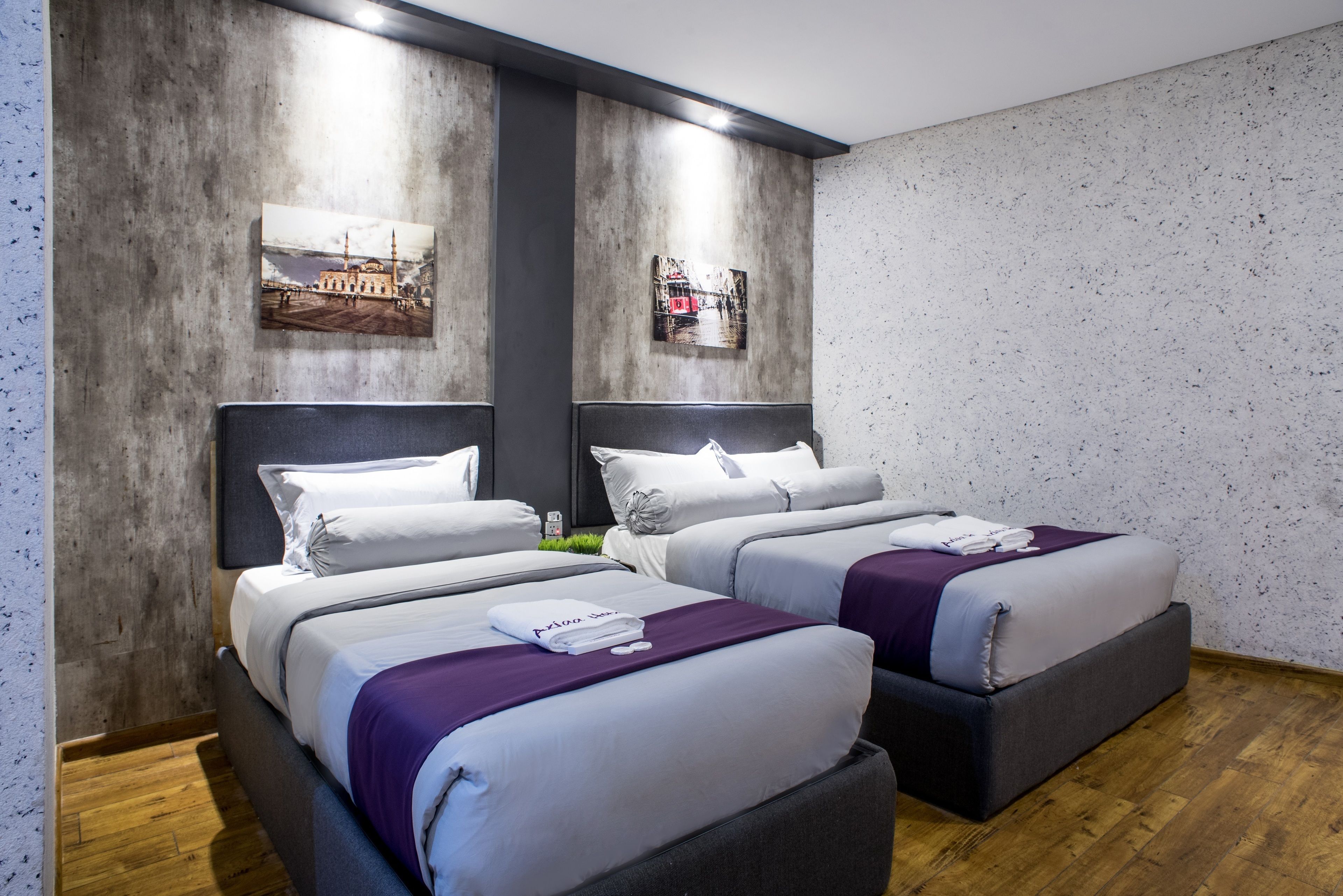 Habitación de hotel con dos camas, cada una con cabecero, suelo de madera y una pared con dos cuadros enmarcados.