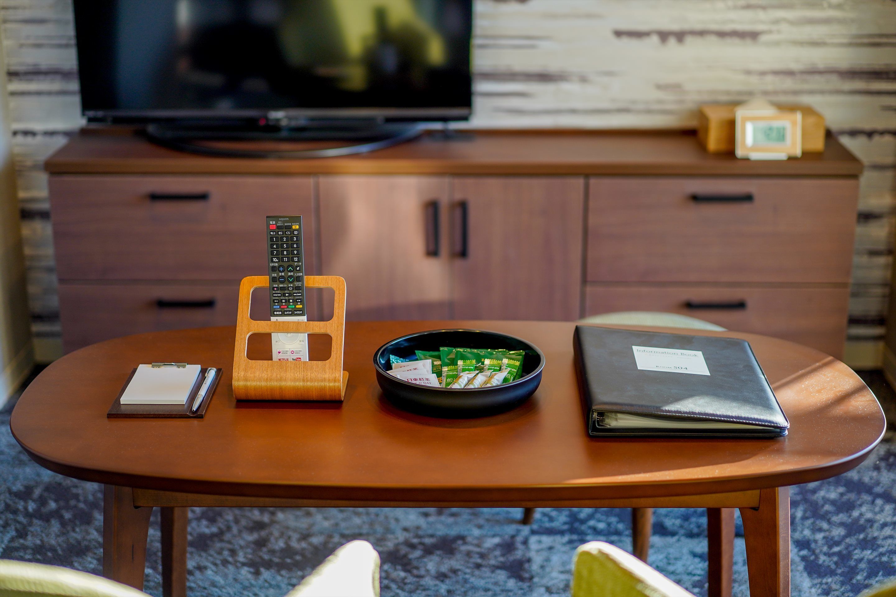 Een houten tafel met een afstandsbediening, een schaal met groene zakjes, een notitieboekje en een gesloten laptop.