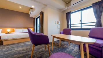 A hotel room with a bed, a desk, purple chairs, and a large window with curtains.