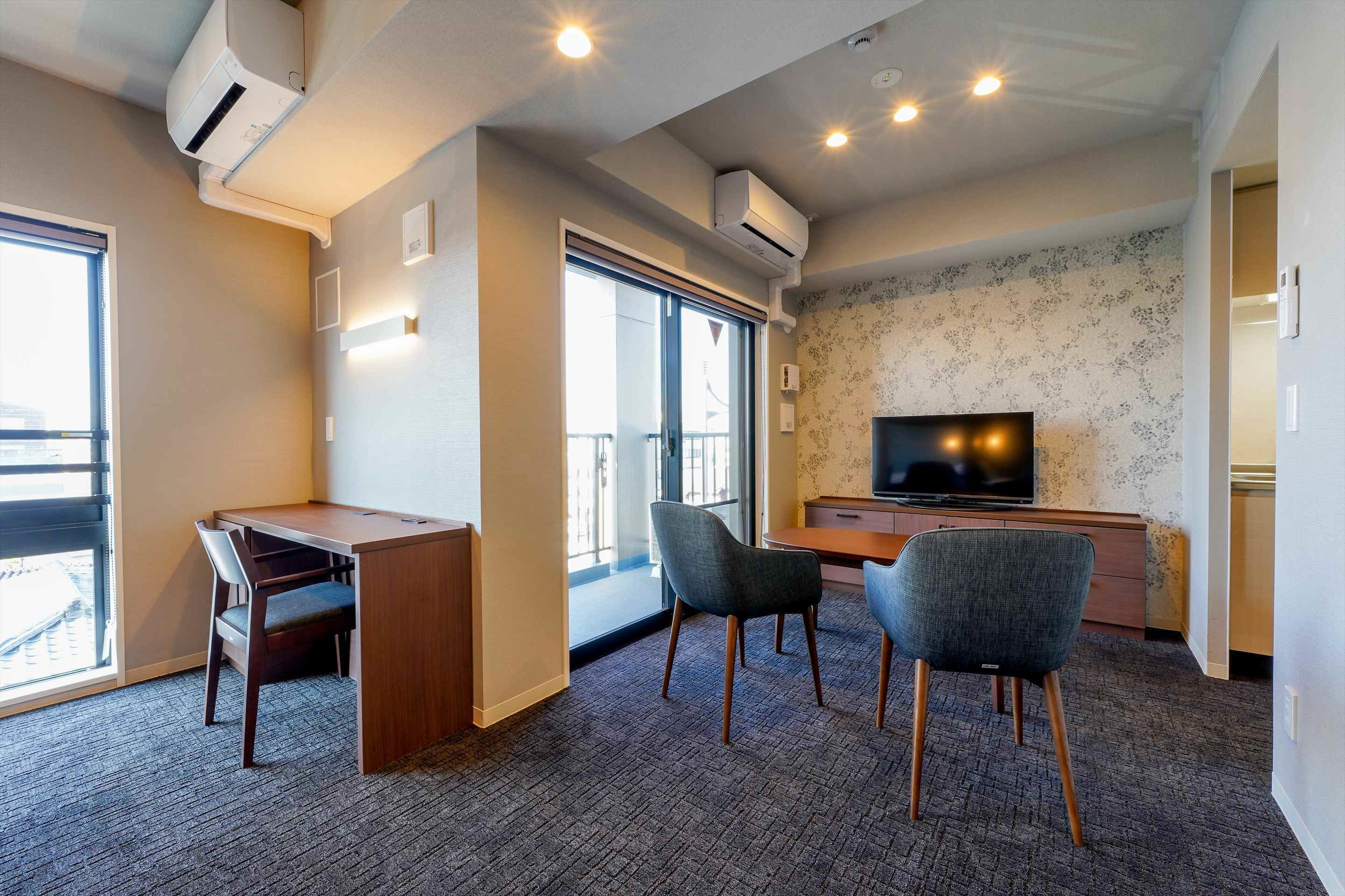 A hotel room with a desk, two chairs, a television, and a large window.