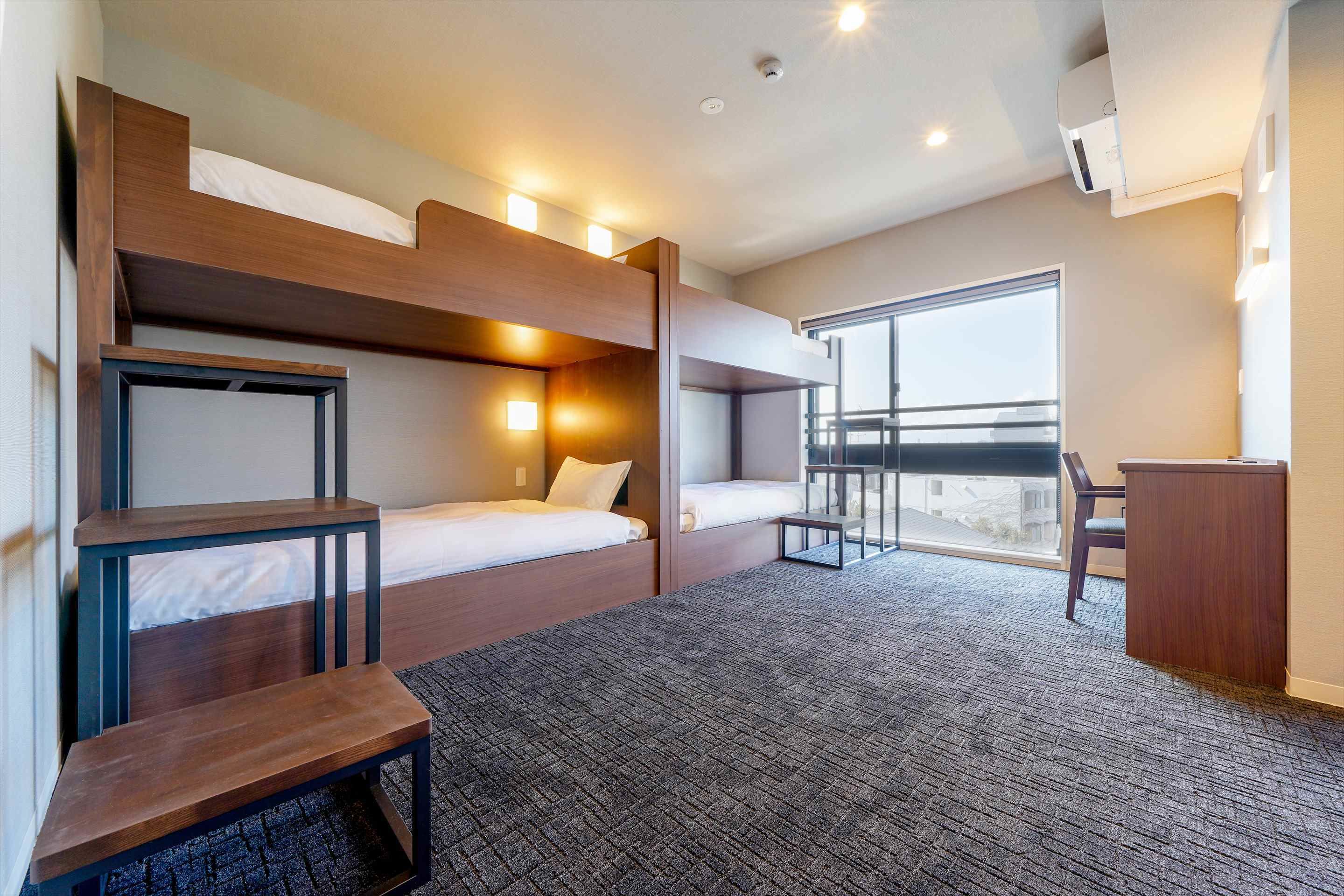 A modern hotel room with bunk beds, a desk, and a large window offering a city view.