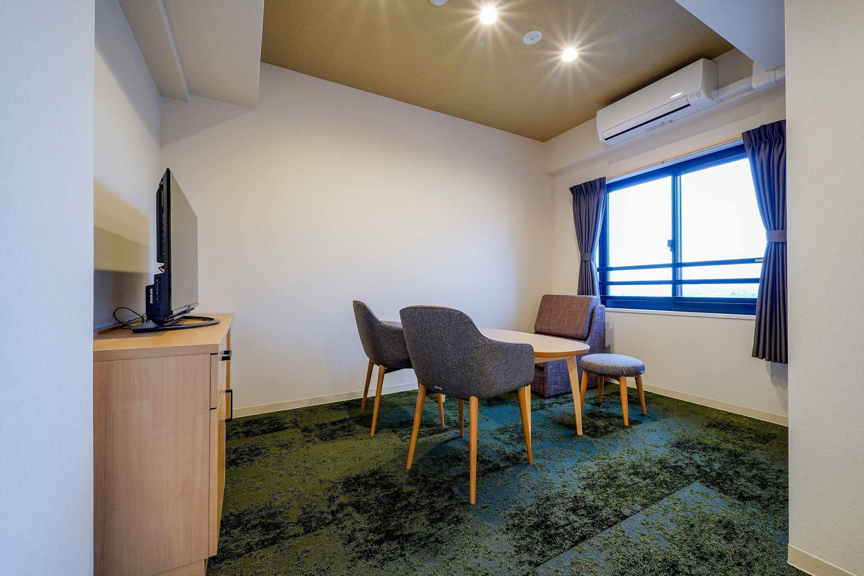 Una habitación pequeña con una mesa de comedor, dos sillas, un televisor en un mueble bajo, una ventana con cortinas y un equipo de aire acondicionado.