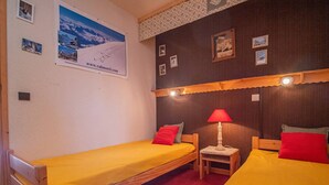 Et værelse med to senge af træ, hver med et gult lagen og røde puder. Der er en lille træstol og et bord med en rød lampe. Væggene er pyntet med billeder i rammer og en stor plakat.