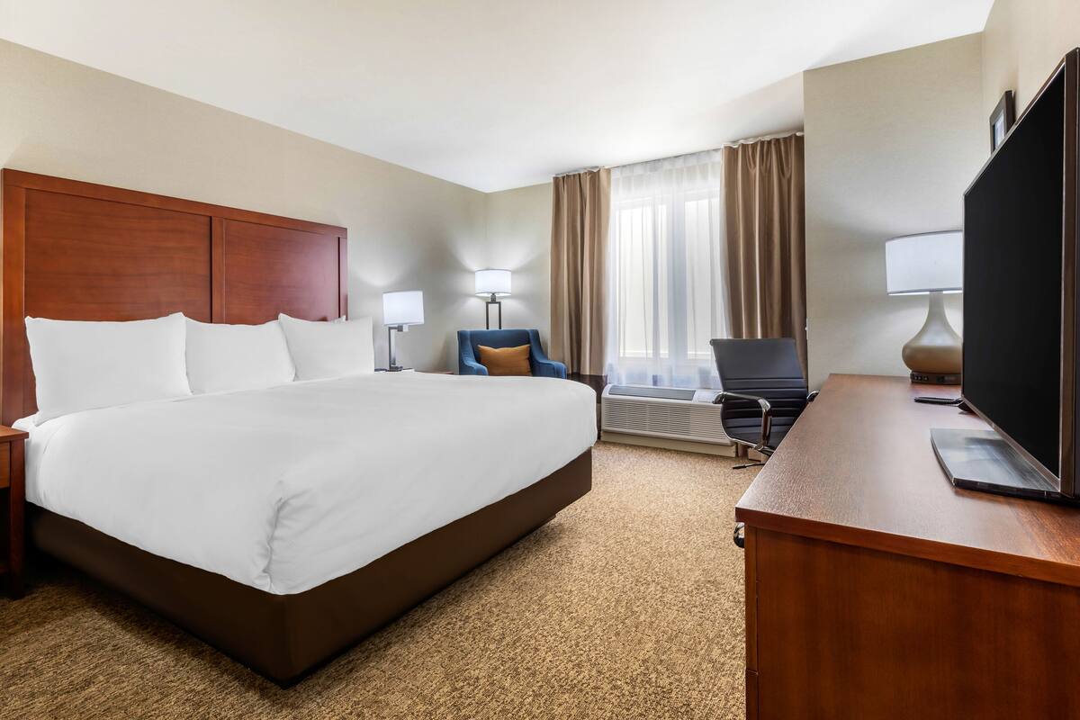 Quarto de hotel com uma cama grande, uma escrivaninha com um laptop, uma cadeira e uma televisão.