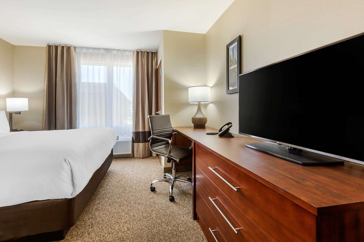 Quarto de hotel com cama, escrivaninha, televisão de tela plana, poltrona, luminária e uma janela grande com cortinas.