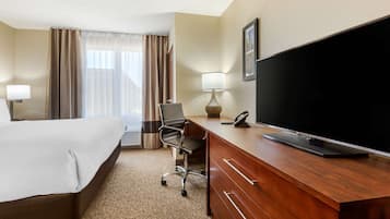 A hotel room with a bed, a desk with a flat-screen TV, a chair, a lamp, and a large window with curtains.
