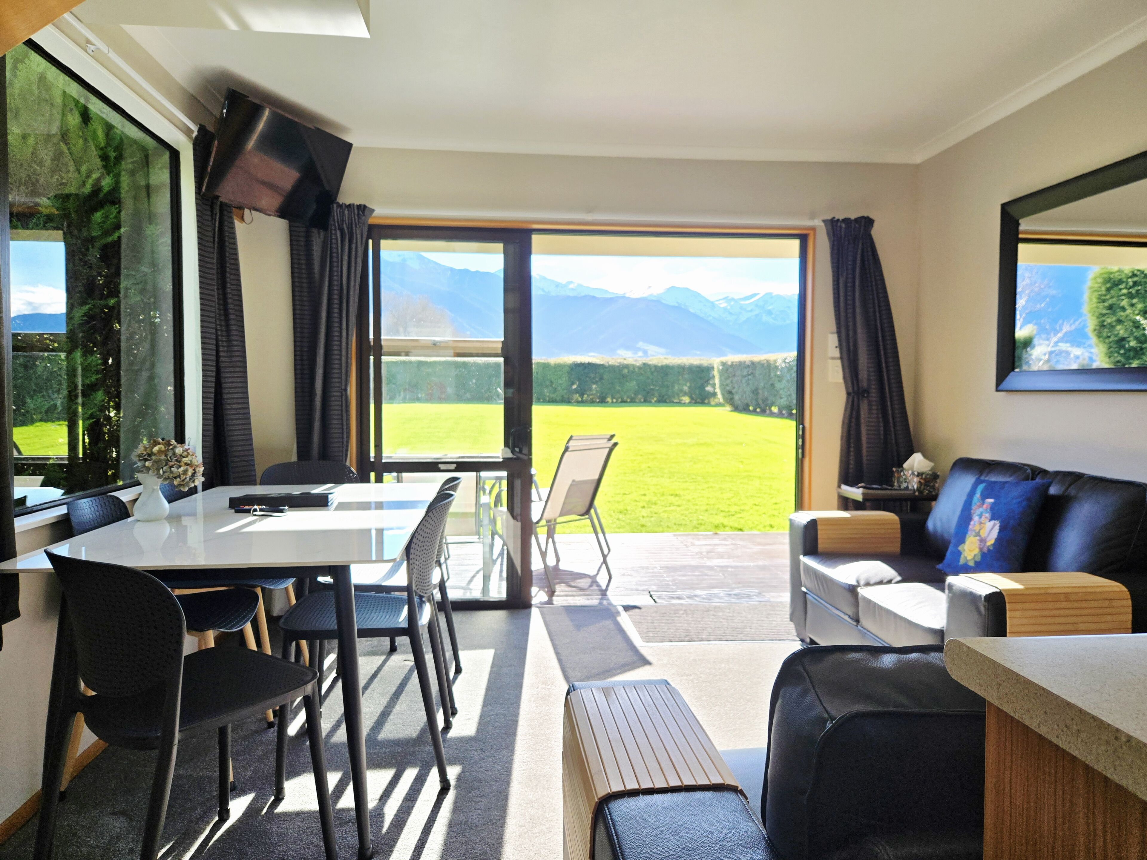 Une pièce comprenant une table à manger, des chaises, un canapé et un téléviseur. Une grande fenêtre offre une vue sur les montagnes et un champ verdoyant.