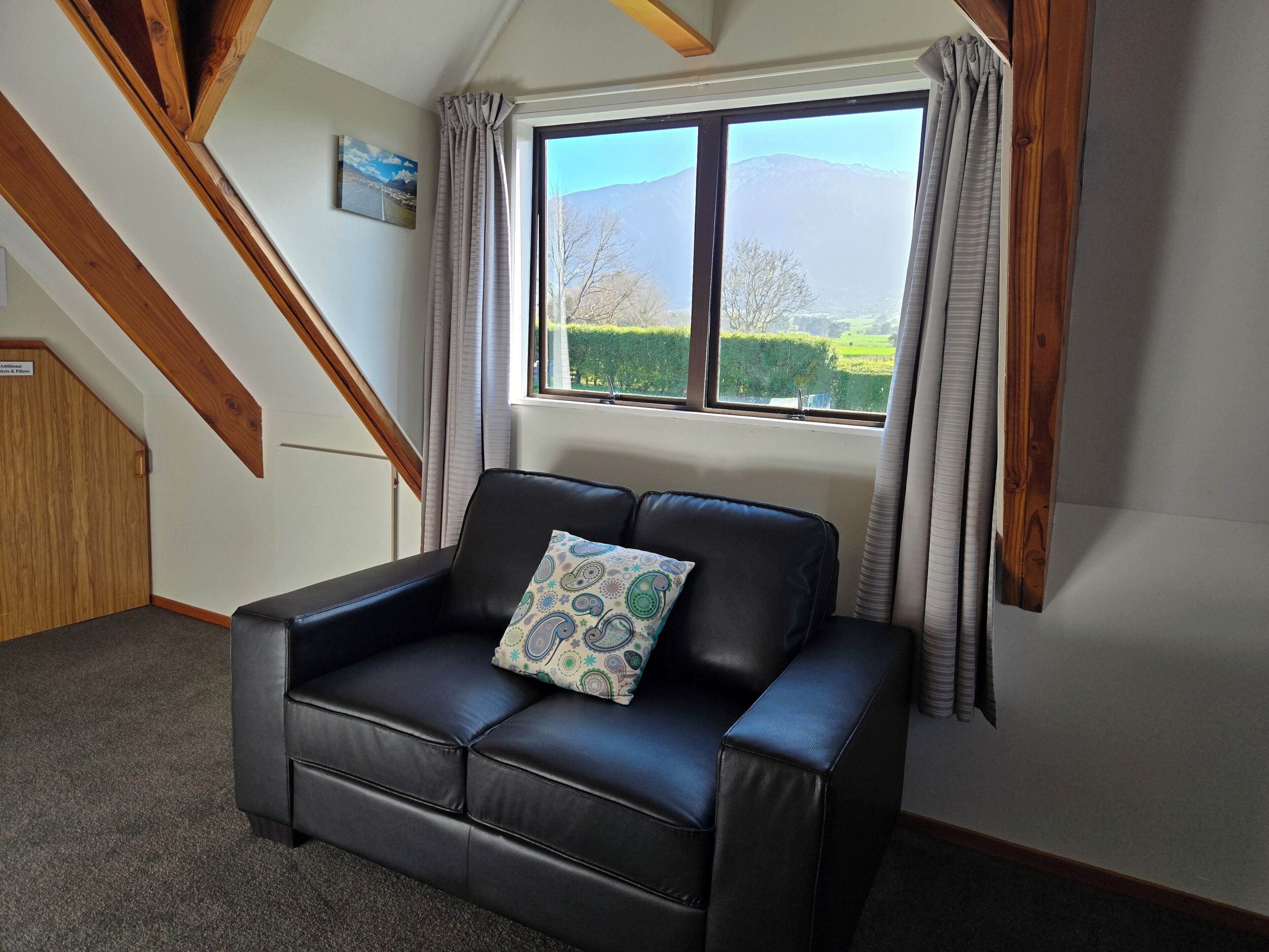 Un sofá de cuero negro con un cojín estampado en una habitación con un gran ventanal, vigas de madera y vistas a la montaña.