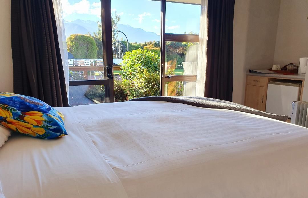 A hotel room with a large bed, a view of a garden and mountains through the window, and a kitchenette area.
