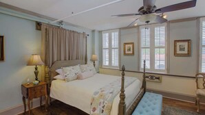 Ein Schlafzimmer mit Himmelbett, Deckenventilator, einer hölzernen Bank und einem Fenster mit Rollläden.