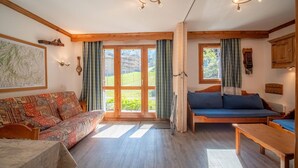 A cozy room with a sofa, a bed, and a wooden bench. There are large windows with curtains, a map on the wall, and a clock. - Divisible studio, near slopes and shops, south terrace (Valmorel)