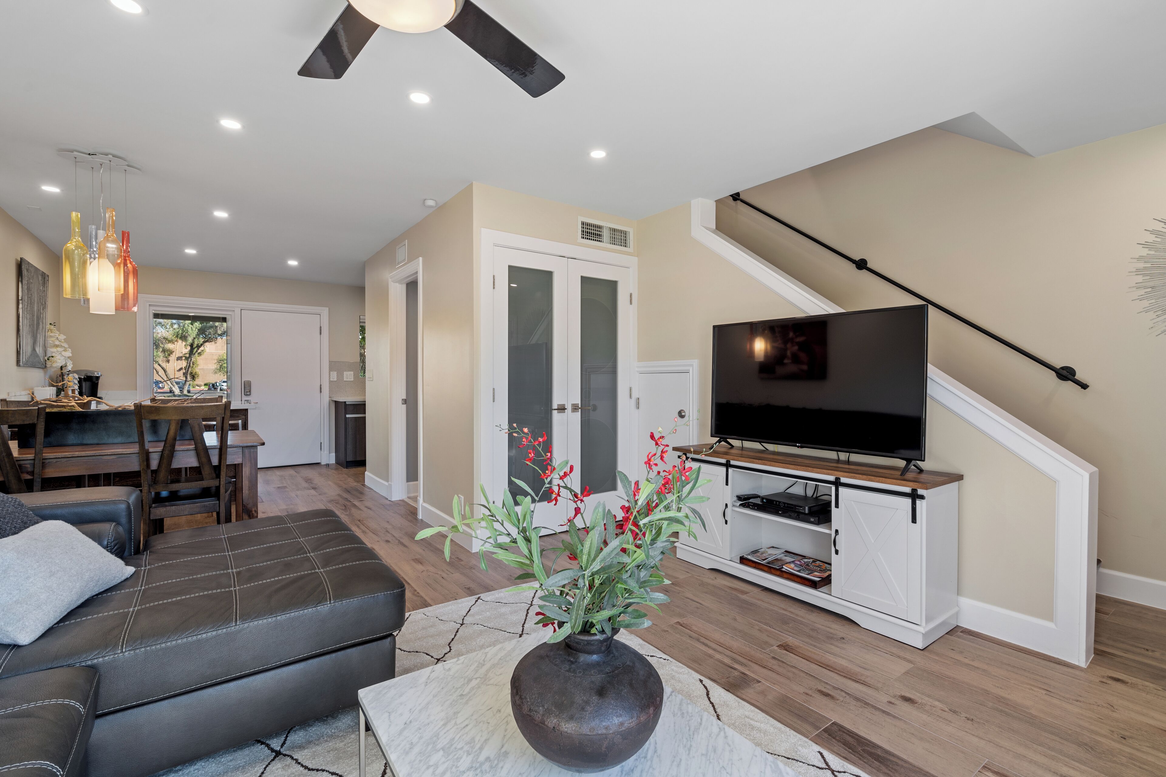 A modern living room with a sofa, a TV on a wooden stand, a vase with flowers, and a dining area with a table and chairs.