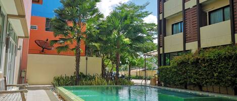 Una piscina con borde de azulejos, rodeada de vegetación y un edificio de colores variados.