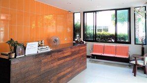 A reception area with a wooden reception desk, orange tiled walls, a large window with a view of greenery, and a bench with a red cushion. - The Wonders Hotel (Chaiyaphum)