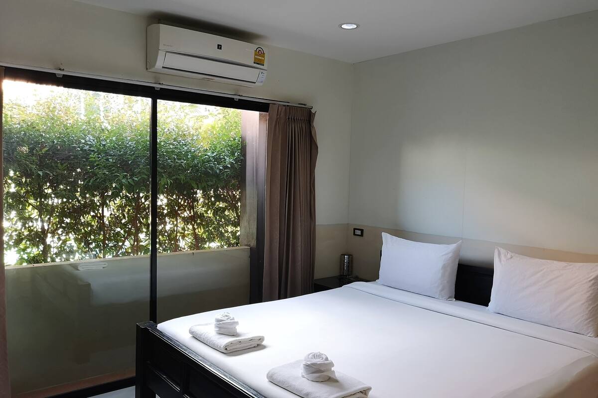 A hotel room with a bed, a window with curtains, an air conditioning unit, and a view of greenery outside.