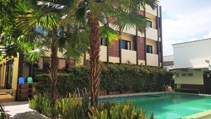 A modern building with a swimming pool, surrounded by lush greenery and palm trees. - The Wonders Hotel (Chaiyaphum)