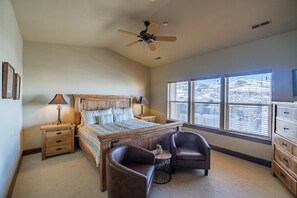Une chambre à coucher avec un grand lit, deux tables de chevet, un ventilateur de plafond, une fenêtre avec des stores et une commode.