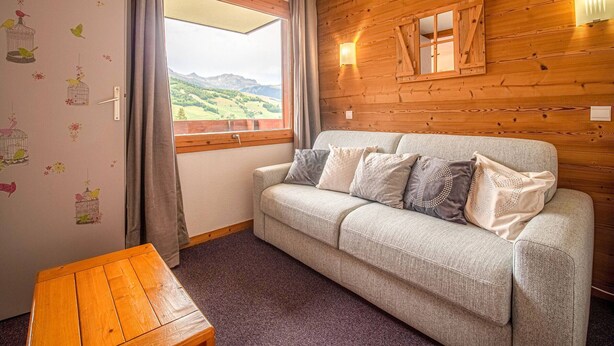 Ein gemütliches Zimmer mit einer Holzwand, einem Sofa mit Kissen, einem Holztisch und einem Fenster mit Blick auf grüne Hügel.