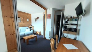 Ein gemütliches Wohnzimmer mit einer Couch, einem Couchtisch und einem Esstisch mit Stühlen. Es gibt eine Küchenzeile mit einer Speisekammer für Küchenutensilien sowie einen Fernseher.