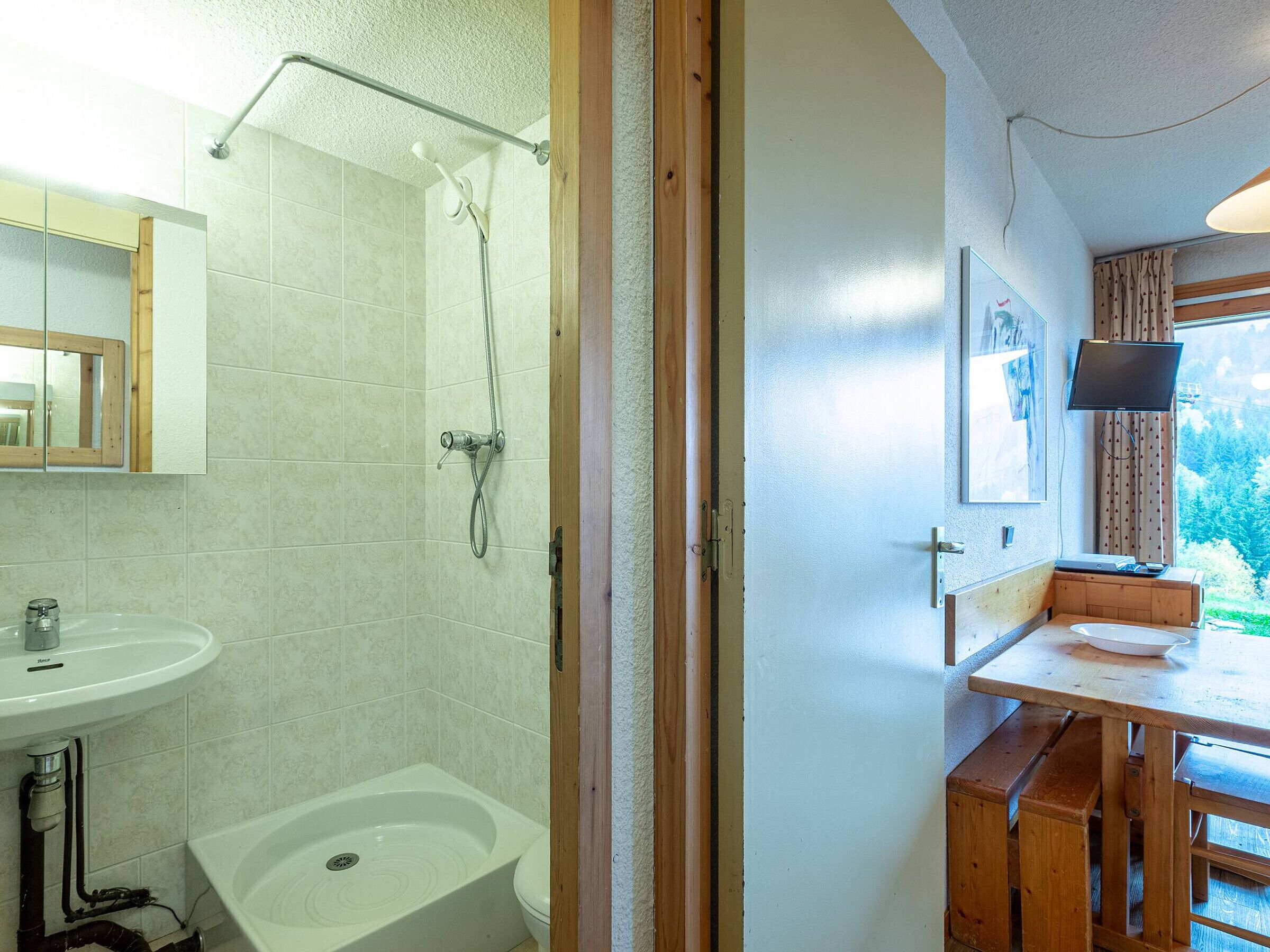 Ein Badezimmer mit Dusche, Waschbecken und Toilette. Es gibt eine hölzerne Bank und ein an der Wand befestigtes Fernsehgerät.