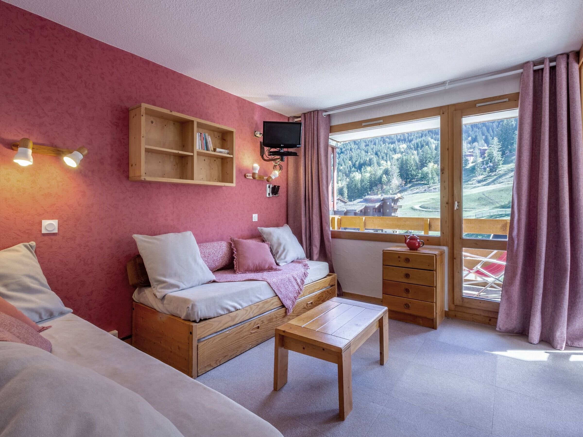 A cozy room with a wooden bed, a wooden table, a TV mounted on the wall, and a window with a view of a forested area.