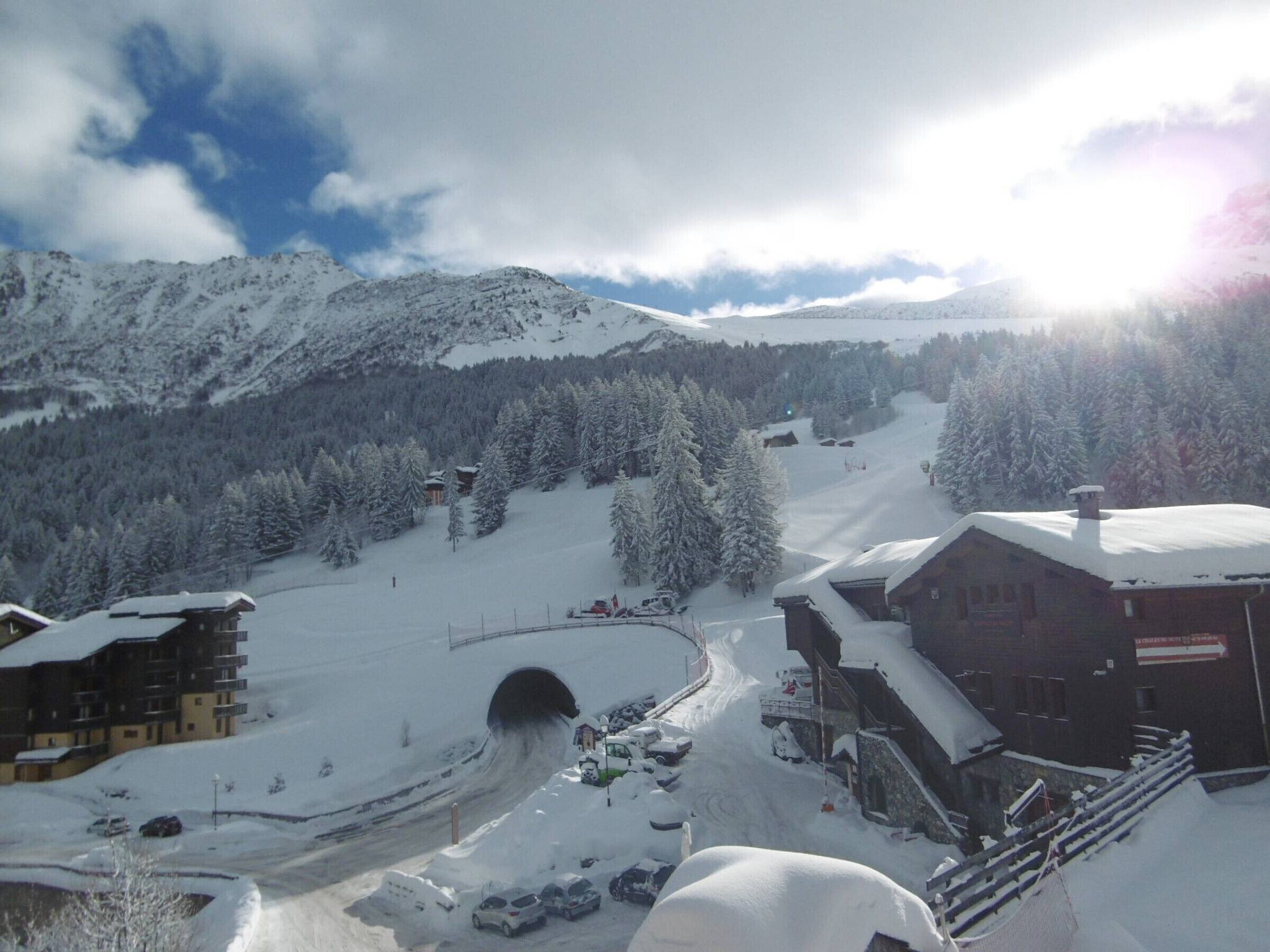 A snow-covered mountain village with a ski lift, buildings, and a road leading to a tunnel.