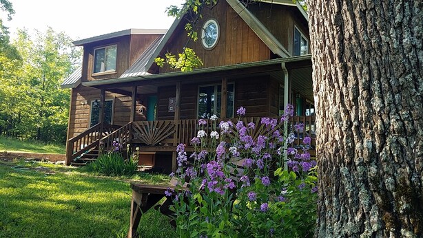 A wooden cabin with a porch, surrounded by greenery and purple flowers. - WIFI!! Big Oak Cabin, ozarks, hot tub, rivers! (Tecumseh)