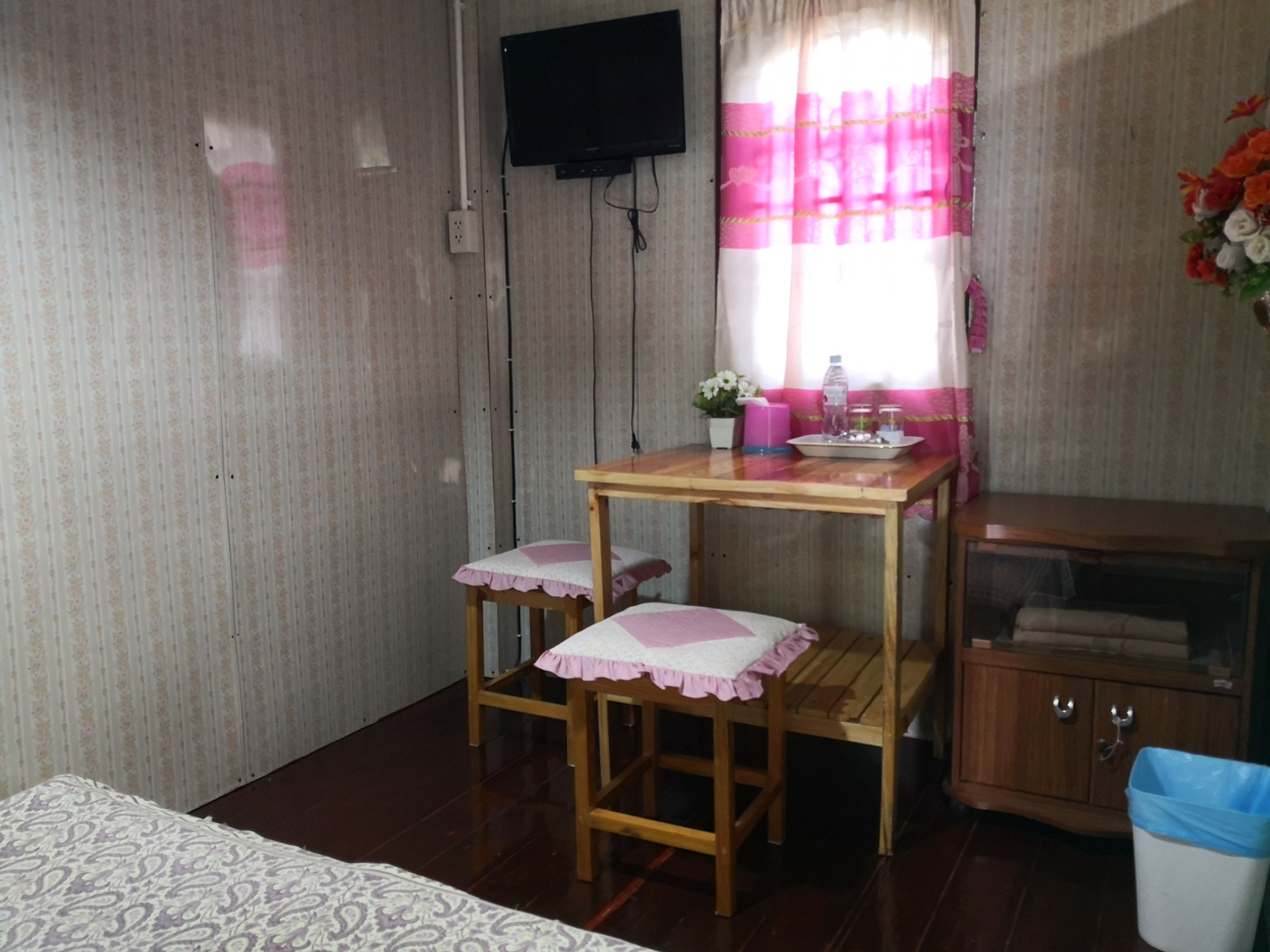 Ein kleines Zimmer mit einem Holztisch und zwei passenden Hocker, einem an der Wand montierten Fernseher, einem Fenster mit rosa Vorhängen und einem Schrank mit blauem Mülleimer.